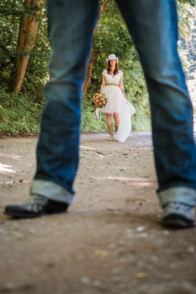 Hochzeitskrone Eine Braut in weißem Kleid und Hut steht auf einem Waldweg und hält Blumen in der Hand, umrahmt von den Beinen einer Person in Jeans und Stiefeln. Ein authentischer Moment, festgehalten von einem Hochzeitsfotografen in Wien und Niederösterreich, der durch seine Linse echte Freude zum Leben erweckt.