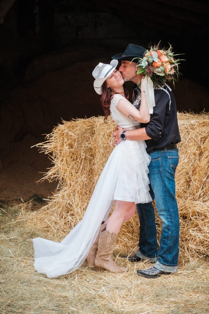 Hochzeitskrone Ein Paar mit Cowboyhüten umarmt sich vor Heuballen, die Frau hält einen Blumenstrauß in der Hand. Sie trägt ein weißes Kleid und Cowboystiefel, er Jeans und ein schwarzes Hemd – dieser Hochzeitsfotograf aus Wien und Niederösterreich fängt den Charme natürlicher Hochzeitsfotos ein.