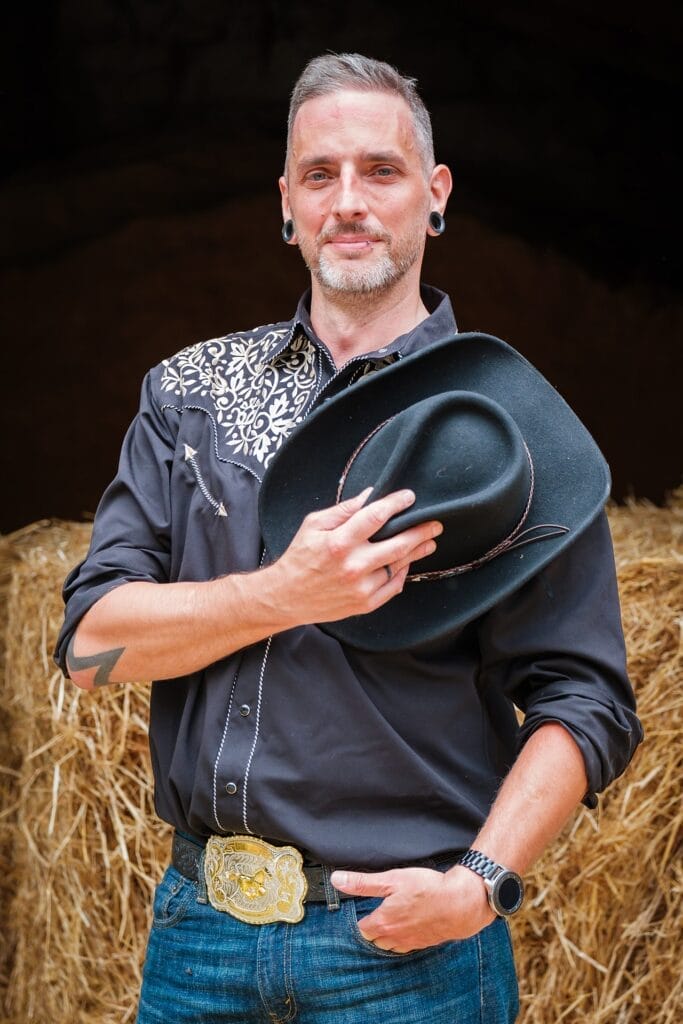 Hochzeitskrone Ein Mann im Westernhemd und Jeans hält einen Cowboyhut und fängt echte Momente vor Heuballen ein.