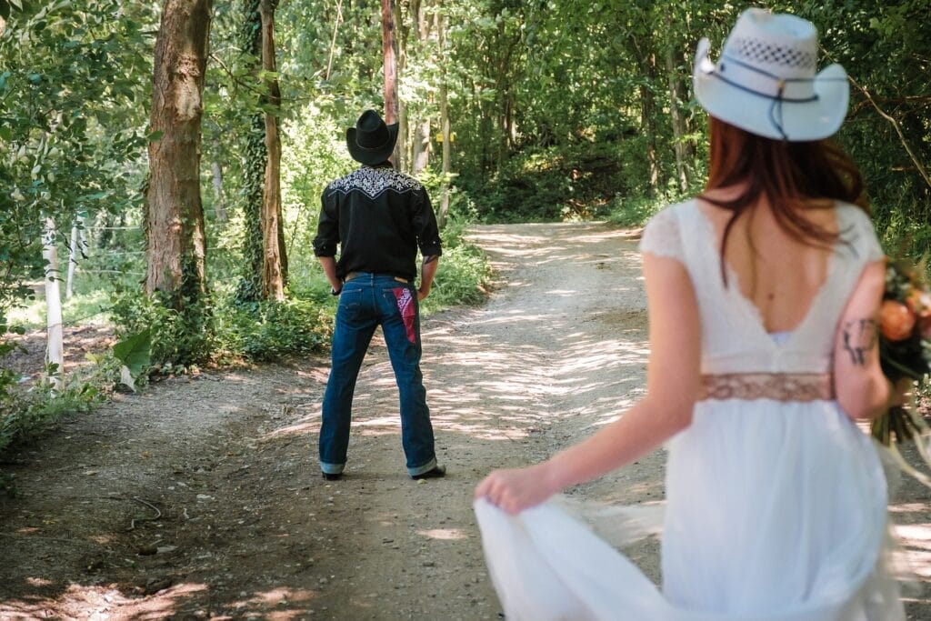 Hochzeitskrone Eine Frau im weißen Kleid nähert sich auf einem Waldweg einem Mann mit Cowboyhut und Jeans und macht natürliche Hochzeitsfotos voller echter Momente.