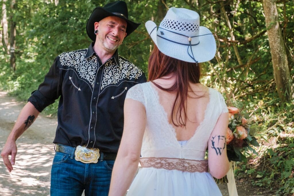 Hochzeitskrone Ein Mann im schwarzen Cowboy-Outfit lächelt eine Frau im weißen Kleid und Cowboyhut auf einem Waldweg an und fängt echte Momente ein, die sich für atemberaubende natürliche Hochzeitsfotos eignen würden.