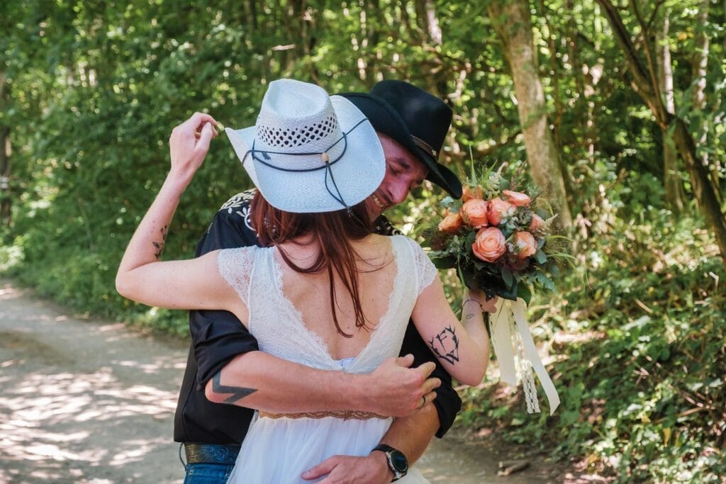 Hochzeitskrone Ein Paar umarmt sich auf einem Waldweg; Die Frau im weißen Kleid und Hut hält einen Blumenstrauß in der Hand, während der Mann ein schwarzes Hemd und einen Hut trägt. Diese echten Momente fangen authentische Hochzeitsfotos von einem Hochzeitsfotografen in Wien und Niederösterreich ein.