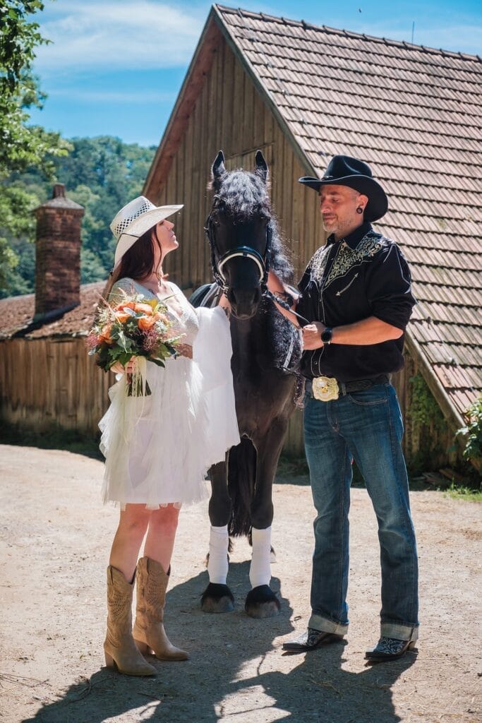 Hochzeitskrone Eine Frau im weißen Kleid und ein Mann im Cowboy-Outfit stehen mit einem schwarzen Pferd da, ihr Blumenstrauß fängt das Sonnenlicht ein. Aufgenommen an einem sonnigen Tag im Freien in der Nähe eines rustikalen Gebäudes, verkörpern diese natürlichen Hochzeitsfotos echte Momente der Freude und Liebe.