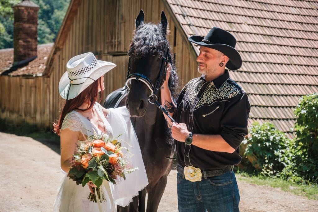 Hochzeitskrone Braut und Bräutigam in Westernkleidung stehen neben einem schwarzen Pferd, wobei die Braut einen Blumenstrauß hält. Sie posieren neben einer Holzkonstruktion, ideal für natürliche Hochzeitsfotos, und fangen ihren authentischen Moment ein.