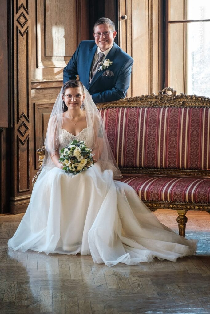 Hochzeitskrone Die Braut im weißen Kleid sitzt mit Blumenstrauß, der Bräutigam im blauen Anzug steht hinter ihr auf einem reich verzierten Sofa in einem von Sonnenlicht durchfluteten Holzraum und fängt bezaubernde Momente ein, die den Sinn für natürliche Hochzeitsfotos charakterisieren.