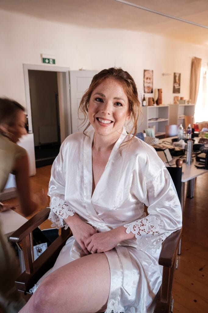 Hochzeitskrone Eine Frau im weißen Morgenmantel sitzt auf einem Stuhl in einem Raum mit Holzboden, lächelt in die Kamera und fängt bezaubernde Momente ein. Eine verschwommene Person bewegt sich im Hintergrund und verleiht den natürlichen Hochzeitsfotos ihren Charme.