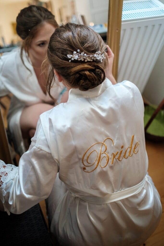 Hochzeitskrone Eine Braut im weißen Gewand mit der Aufschrift „Braut“ sitzt vor einem Spiegel, ihr Haar ist mit einer dekorativen Spange frisiert, und fängt bezaubernde Momente voller Eleganz und Freude ein – wahrhaft natürliche Hochzeitsfotografie in Niederösterreich.