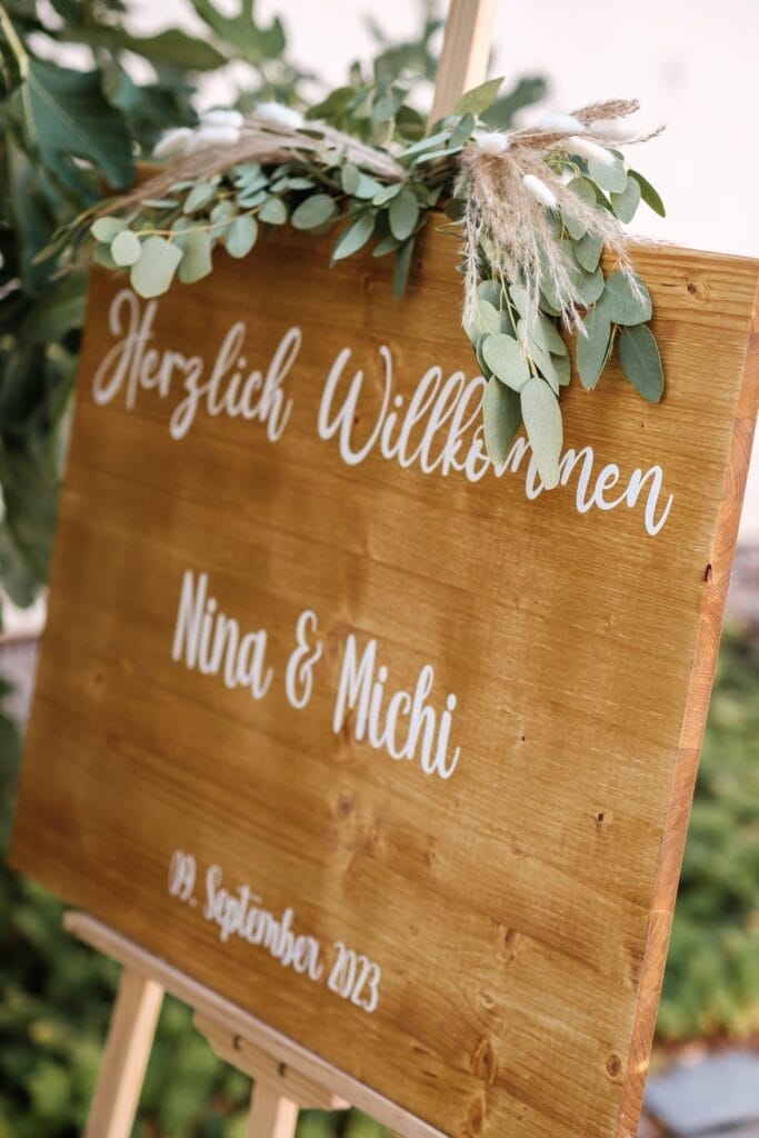 Hochzeitskrone Ein mit Grün geschmücktes hölzernes Willkommensschild mit der herzlichen Aufschrift „Herzlich Willkommen Nina & Michi 19. September 2023“ lädt die Gäste ein, bezaubernde Momente festzuhalten. Dieser Rahmen ist perfekt für natürliche Hochzeitsfotos und verkörpert die Essenz der Hochzeitsfotografie in Niederösterreich.