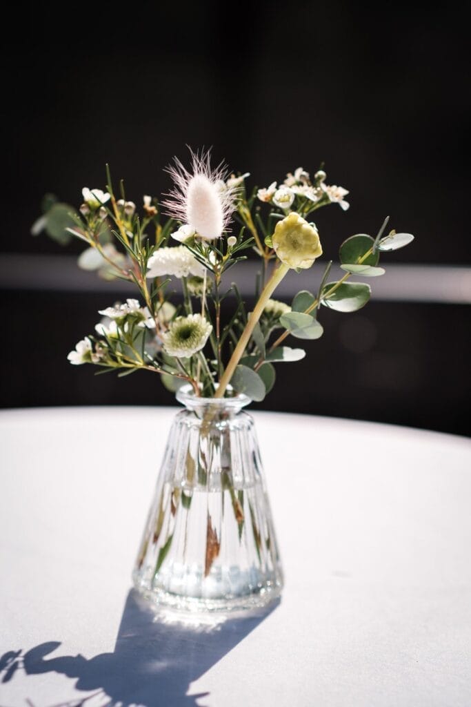 Hochzeitskrone Ein kleiner Strauß verschiedener Blumen in einer klaren Glasvase auf einem weißen Tisch, der bezaubernde Momente vor einem dunklen Hintergrund einfängt und an natürliche Hochzeitsfotografie in Niederösterreich erinnert.