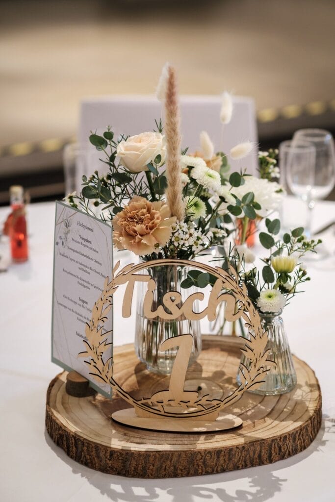 Hochzeitskrone Tischdekoration aus Holz mit dem Schild „Tisch 1“, umgeben von kleinen Glasvasen mit weißen und pfirsichfarbenen Blumen, einer Menükarte und Weingläsern auf einer makellos weißen Tischdecke – perfekt, um bezaubernde Momente in der Hochzeitsfotografie in Niederösterreich festzuhalten.