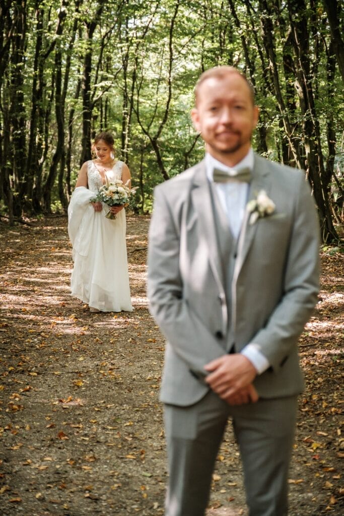 Hochzeitskrone Ein Bräutigam im grauen Anzug steht scharf auf einem Waldweg und fängt bezaubernde Momente ein, während sich die Braut im weißen Kleid von hinten, leicht verschwommen, nähert – eine perfekte Szene natürlicher Hochzeitsfotos, die Hochzeitsfotografie in Niederösterreich zeigt.