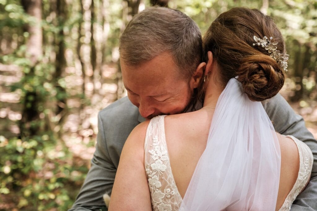 Hochzeitskrone In einer Waldkulisse umarmt ein Bräutigam im Anzug herzlich seine Braut, die in ihrem Spitzenkleid und Schleier strahlt. Diese bezaubernden Momente fangen die Essenz natürlicher Hochzeitsfotos ein und schaffen Erinnerungen, die man für immer schätzen wird.