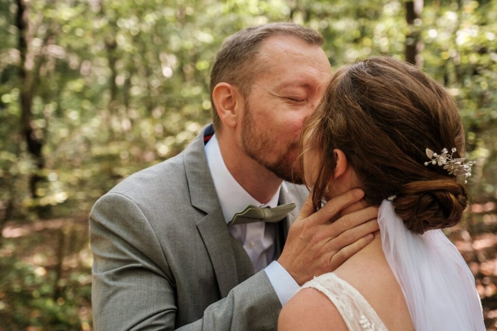 Hochzeitskrone In einer ruhigen Waldkulisse küsst ein Mann im Anzug eine Frau zärtlich auf die Stirn. Ihr Haar ist zu einer eleganten Hochsteckfrisur gestylt, geschmückt mit einer dekorativen Nadel und einem Schleier. Bezaubernde Momente der Hochzeitsfotografie in Niederösterreich werden hier festgehalten.