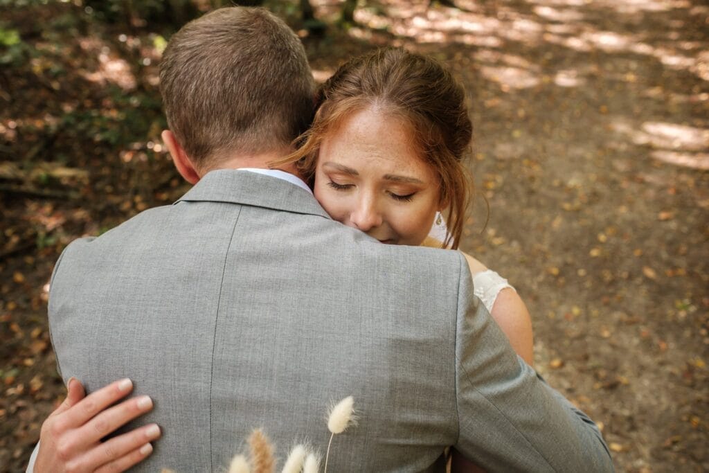 Hochzeitskrone Auf einem Waldweg umarmt eine Frau im weißen Kleid einen Mann im grauen Anzug, eingefangen inmitten bezaubernder Momente.