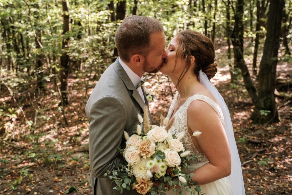 Hochzeitskrone In einem sonnenbeschienenen Wald küssen sich Braut und Bräutigam und schaffen bezaubernde Momente. Sie hält einen Strauß weißer und blassrosa Blumen in den Händen und fängt in dieser bezaubernden Szene die Essenz natürlicher Hochzeitsfotos ein.