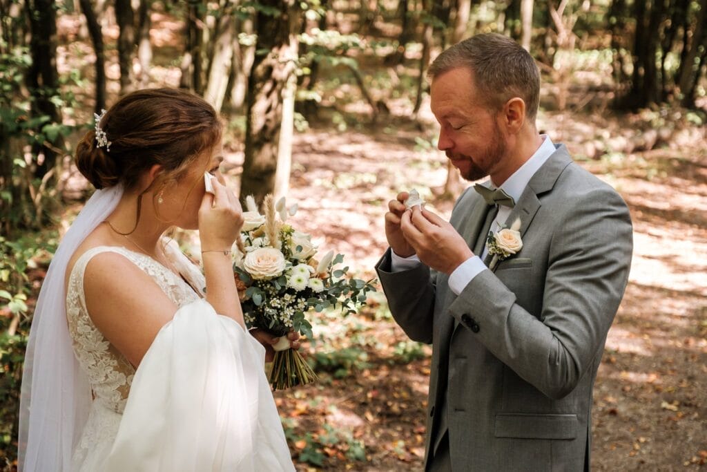 Hochzeitskrone In einem Waldstück teilen Braut und Bräutigam bezaubernde Momente und wischen sich dabei Tränen ab. Die Braut hält einen Strauß weißer Blumen in der Hand und ihr Spitzenkleid ist entzückend; der Bräutigam trägt einen grauen Anzug mit einer Boutonniere – ein Beispiel für natürliche Hochzeitsfotos in Hochezitsfotografie in Niederösterreich.