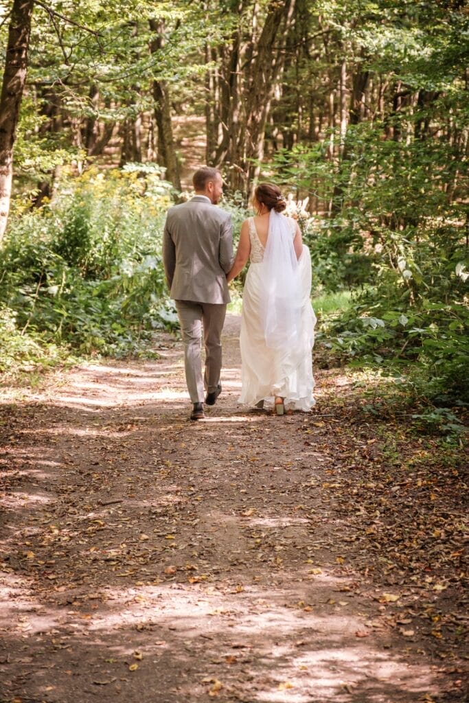 Hochzeitskrone Ein Paar im Hochzeitskleid geht einen Waldweg entlang und fängt bezaubernde Momente inmitten des üppigen Grüns und des durch die Bäume fallenden Sonnenlichts ein.