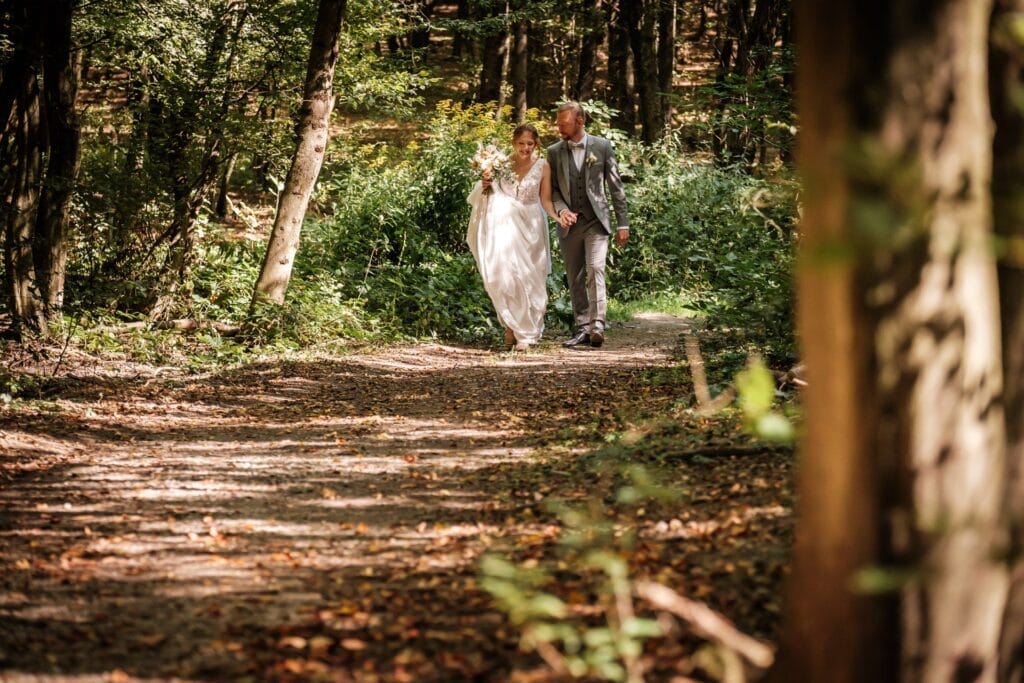 Hochzeitskrone Braut und Bräutigam schlendern einen Waldweg entlang, umgeben von Bäumen und gesprenkeltem Sonnenlicht, und halten bezaubernde Momente natürlicher Hochzeitsfotos fest.