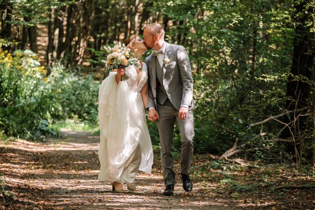 Hochzeitskrone Braut und Bräutigam gehen Hand in Hand einen Waldweg entlang und halten bezaubernde Momente fest. Die Braut hält einen Blumenstrauß, während der Bräutigam einen grauen Anzug trägt, perfekt für natürliche Hochzeitsfotos.