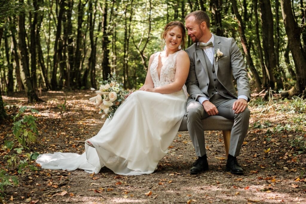 Hochzeitskrone Braut im weißen Kleid und Bräutigam im grauen Anzug sitzen lächelnd auf einer Bank im Wald. Die Braut hält einen Blumenstrauß in der Hand und fängt bezaubernde Momente natürlicher Hochzeitsfotos ein.