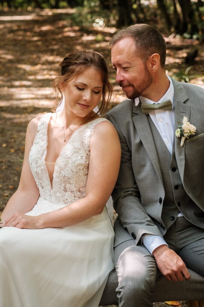 Hochzeitskrone Eine Braut im Spitzenkleid und ein Bräutigam im grauen Anzug sitzen eng aneinandergedrängt auf einer Bank im Freien, umgeben von Bäumen, und halten bezaubernde Momente in natürlichen Hochzeitsfotos fest.