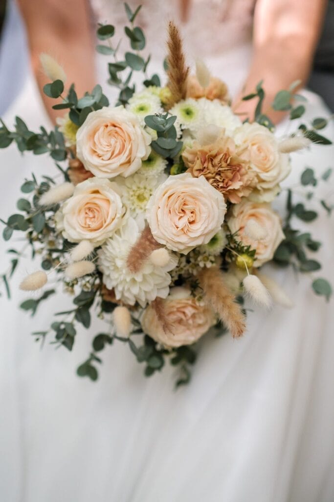 Hochzeitskrone Ein Strauß cremefarbener Rosen, weißer Chrysanthemen, getrockneter Gräser und Eukalyptusblätter, gehalten von einer Person im weißen Kleid, fängt bezaubernde Momente ein.