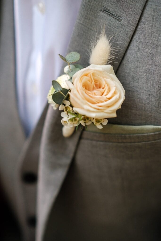 Hochzeitskrone Eine Nahaufnahme einer Ansteckblume an einer grauen Anzugjacke fängt bezaubernde Momente ein, mit einer cremefarbenen Rose, kleinen weißen Blüten, Eukalyptusblättern und einem flauschigen Akzent. Ideal für natürliche Hochzeitsfotos, die die Eleganz der Hochzeitsfotografie in Niederösterreich hervorheben.