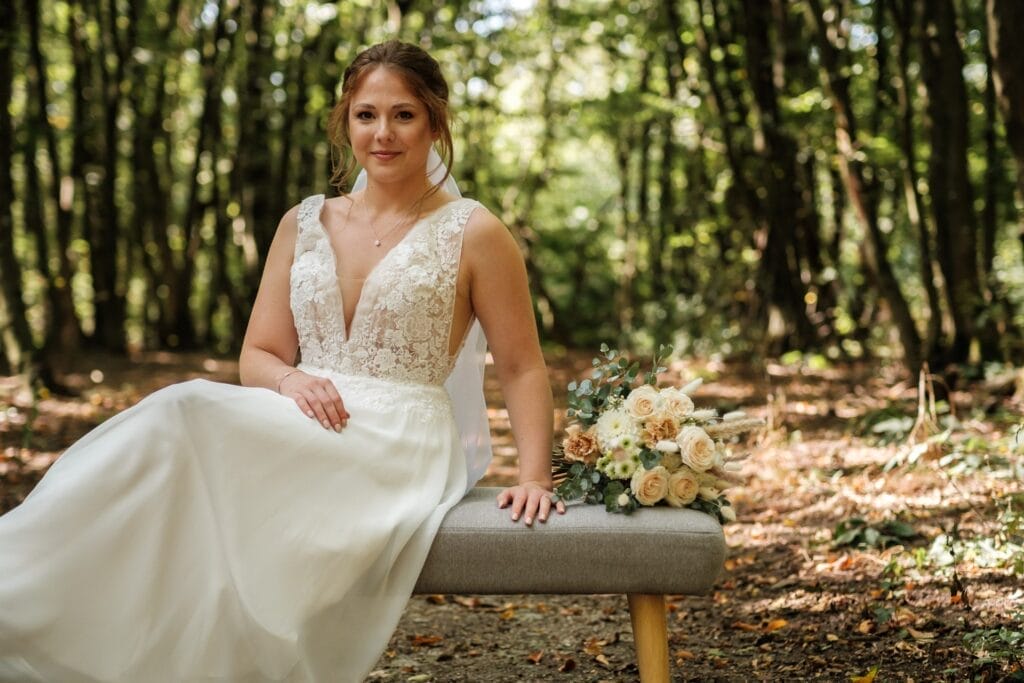 Hochzeitskrone In einer ruhigen Waldlandschaft sitzt eine Braut in einem weißen Spitzenbrautkleid anmutig auf einer Bank und fängt bezaubernde Momente ein. Neben ihr liegt ein Strauß blasser Blumen, der die Essenz natürlicher Hochzeitsfotos verkörpert.