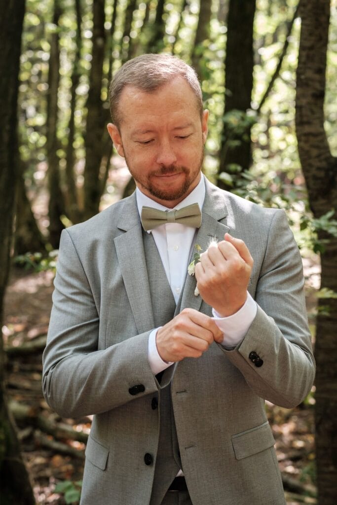 Hochzeitskrone Ein Mann in einem hellgrauen Anzug rückt seine Manschettenknöpfe in einem Waldgebiet zurecht, wobei Bäume und gesprenkeltes Sonnenlicht bezaubernde Momente schaffen. Ideal für natürliche Hochzeitsfotos, fängt diese Szene die Essenz der Hochzeitsfotografie in Niederösterreich ein.