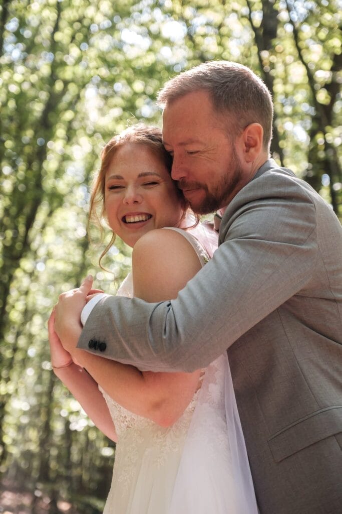 Hochzeitskrone In einem sonnenbeschienenen Wald umarmt sich ein Paar und lächelt, um bezaubernde Momente festzuhalten. Die Frau trägt ein weißes Kleid und der Mann einen grauen Anzug – perfekt für natürliche Hochzeitsfotos in Niederösterreich.