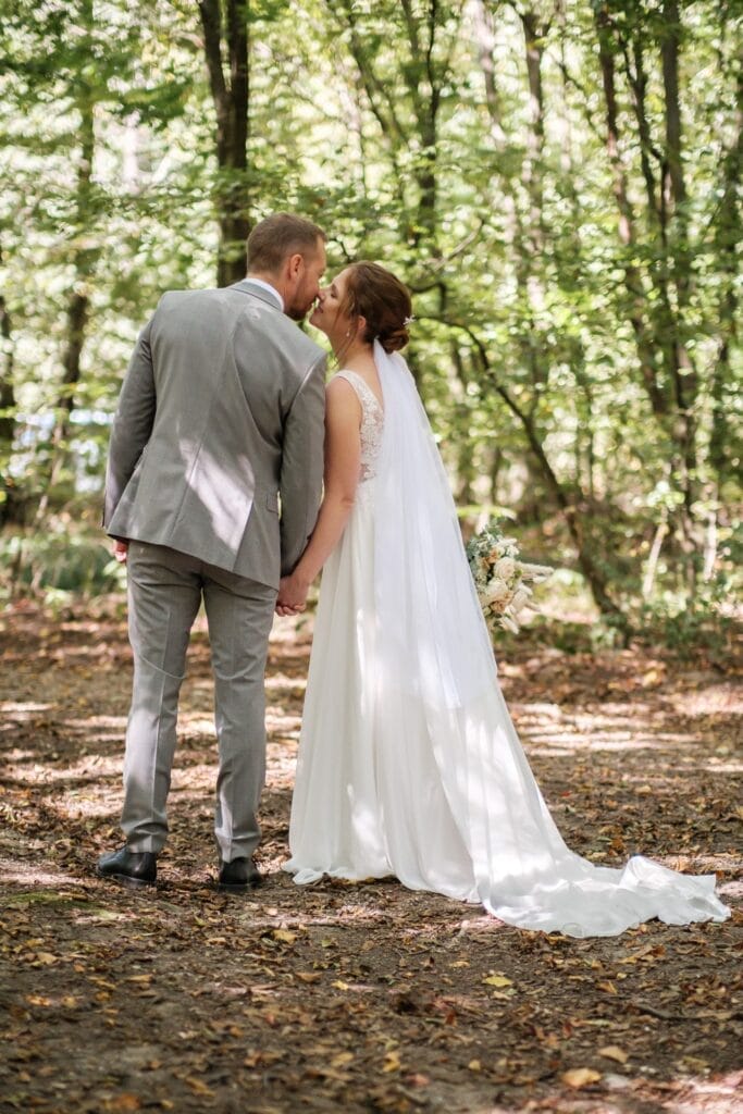 Hochzeitskrone Ein Paar im Hochzeitskleid steht Händchen haltend in einem Waldstück, die Braut in einem weißen Kleid und Schleier, der Bräutigam in einem grauen Anzug. Sie beugen sich zu einem Kuss vor und fangen bezaubernde Momente natürlicher Hochzeitsfotos vor der zauberhaften Kulisse Niederösterreichs ein.