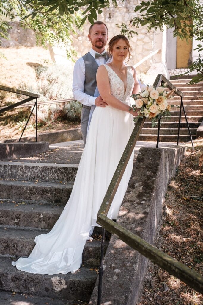 Hochzeitskrone Ein Paar posiert auf Steinstufen im Freien; die Frau im weißen Kleid hält einen Blumenstrauß und verströmt die Stimmung natürlicher Hochzeitsfotos, während der Mann in Hemd und Weste hinter ihr steht. Es geht darum, bezaubernde Momente festzuhalten.