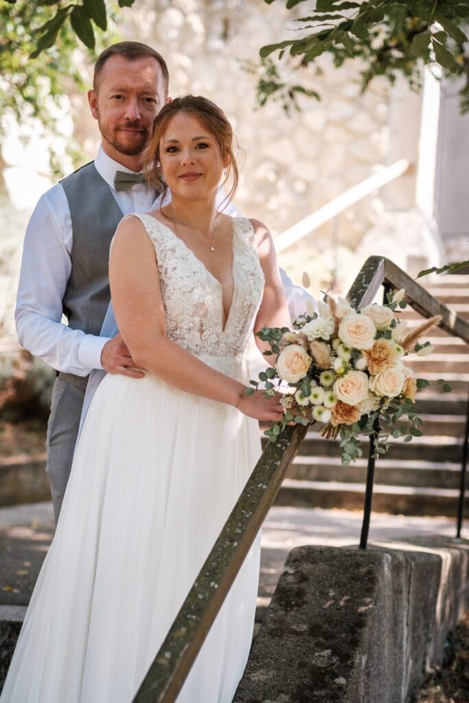 Hochzeitskrone Eine Braut im weißen Kleid und ein Bräutigam in einer grauen Weste posieren an einem Steingeländer und halten bezaubernde Momente mit einem Blumenstrauß fest. Diese natürliche Hochzeitsfotokulisse veranschaulicht die Eleganz der Hochzeitsfotografie in Niederösterreich.