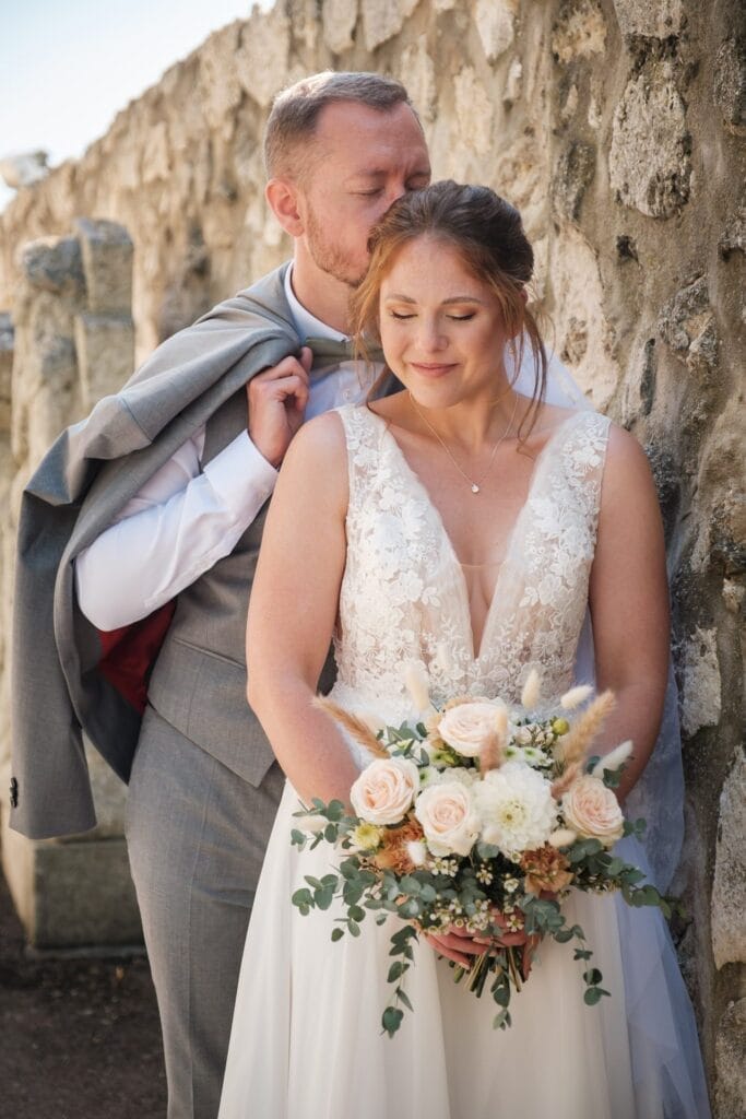 Hochzeitskrone Braut und Bräutigam posieren neben einer Steinmauer und halten bezaubernde Momente fest. Der Bräutigam legt eine Jacke über die Schulter und lehnt sich vor, während die Braut einen Blumenstrauß hält und sanft lächelt. Dies sind natürliche Hochzeitsfotos in Niederösterreich.