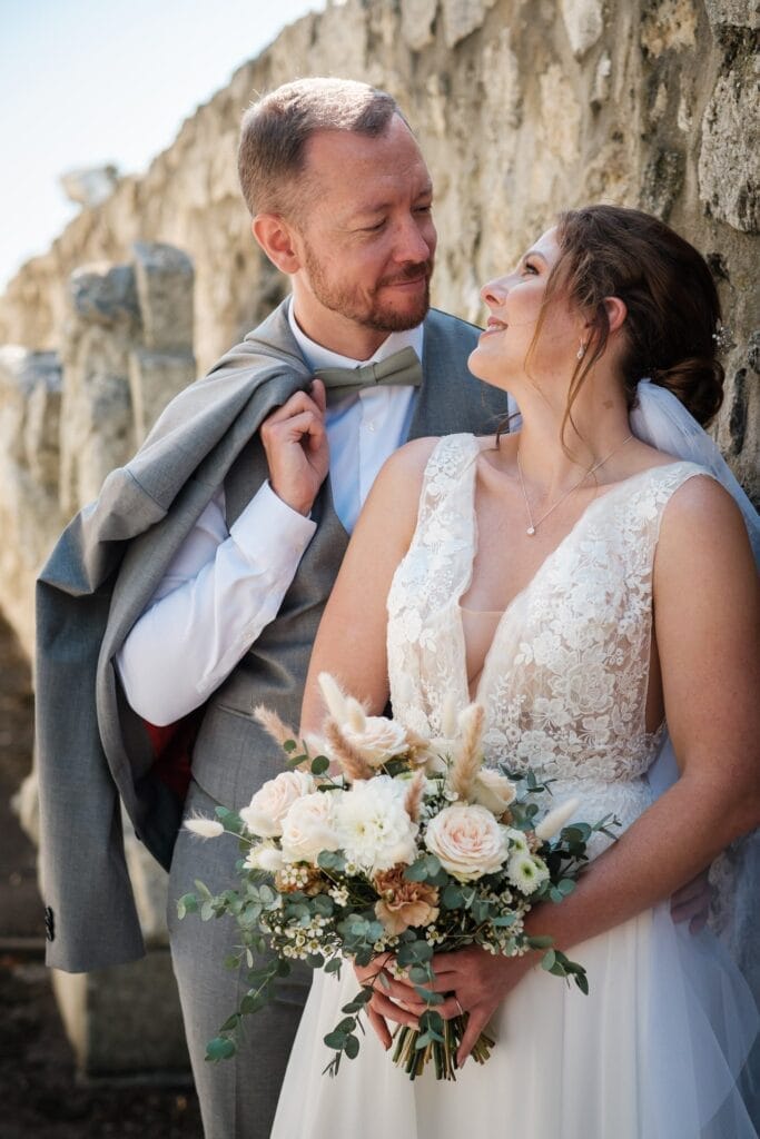 Hochzeitskrone Ein Paar im Hochzeitskleid posiert neben einer Steinmauer und verkörpert natürliche Hochzeitsfotos. Der Bräutigam hängt seine Jacke über die Schulter, während die Braut einen Strauß Rosen und Grünzeug hält. Beide lächeln sich an und fangen bezaubernde Momente ein.