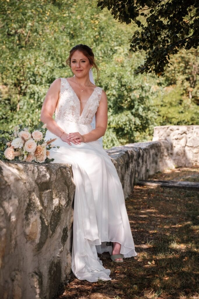 Hochzeitskrone Eine Braut im weißen Brautkleid sitzt auf einer Steinmauer, ihr Blumenstrauß liegt daneben, umgeben von üppigem Grün. Bezaubernde Momente, festgehalten durch natürliche Hochzeitsfotos, bringen den Zauber der Hochzeitsfotografie in Niederösterreich zum Vorschein.