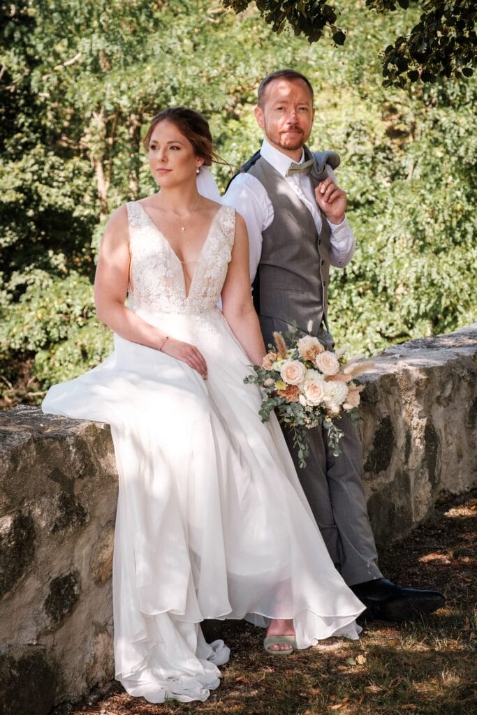 Hochzeitskrone Eine Braut im weißen Kleid mit einem Blumenstrauß in der Hand steht neben einem Bräutigam in Weste und Krawatte und fängt bezaubernde Momente vor der Kulisse einer Steinmauer und von Bäumen ein und verkörpert so natürliche Hochzeitsfotos.