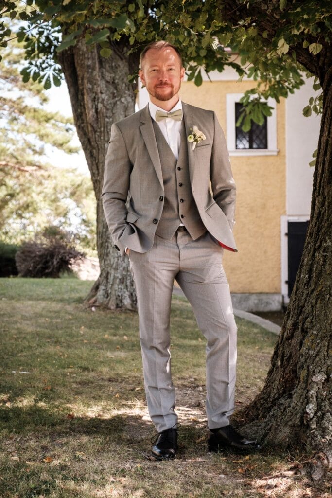 Hochzeitskrone Ein Mann in grauem Anzug und Fliege steht lächelnd unter einem Baum im Freien, die Hände in den Taschen, und fängt bezaubernde Momente der Hochzeitsfotografie in Niederösterreich ein. Im Hintergrund ist ein Gebäude zu sehen, das dieser Naturszene Charme verleiht.