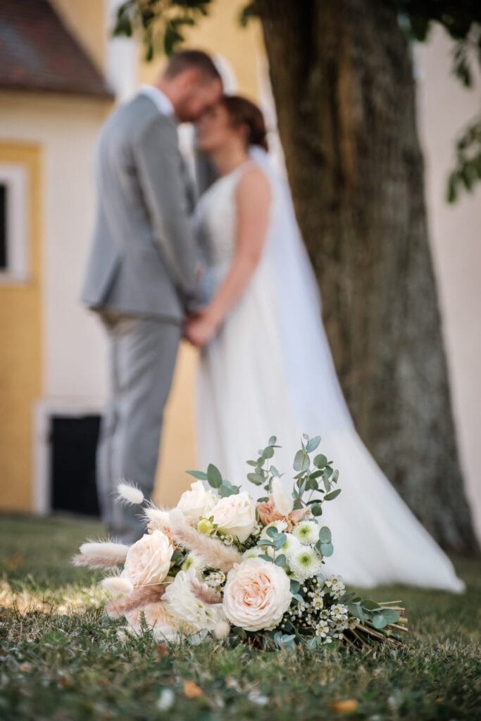 Hochzeitskrone Ein Brautstrauß liegt auf dem Gras und fängt bezaubernde Momente ein, während Braut und Bräutigam im Hintergrund stehen, einander an den Händen halten und gegenüberstehen.