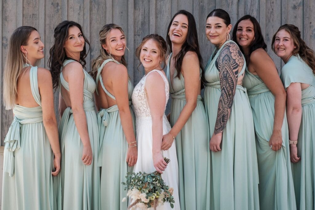 Hochzeitskrone Eine Braut im weißen Kleid posiert mit Brautjungfern in passenden hellgrünen Kleidern vor einer Holzwand. Sie lächeln alle und fangen bezaubernde Momente ein, die natürliche Hochzeitsfotos hervorheben.