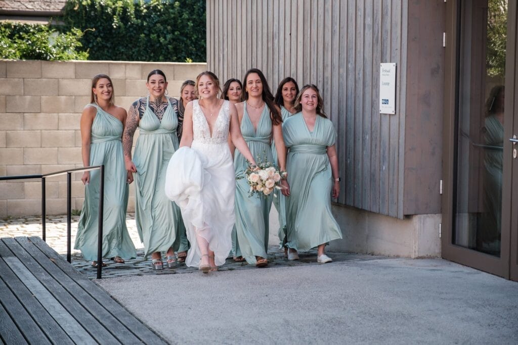 Hochzeitskrone Eine Braut in einem weißen Kleid und Schleier geht mit ihren Brautjungfern in passenden grünen Kleidern vor einem Gebäude spazieren, hält einen Blumenstrauß in der Hand und lächelt inmitten bezaubernder Momente natürlicher Hochzeitsfotos.