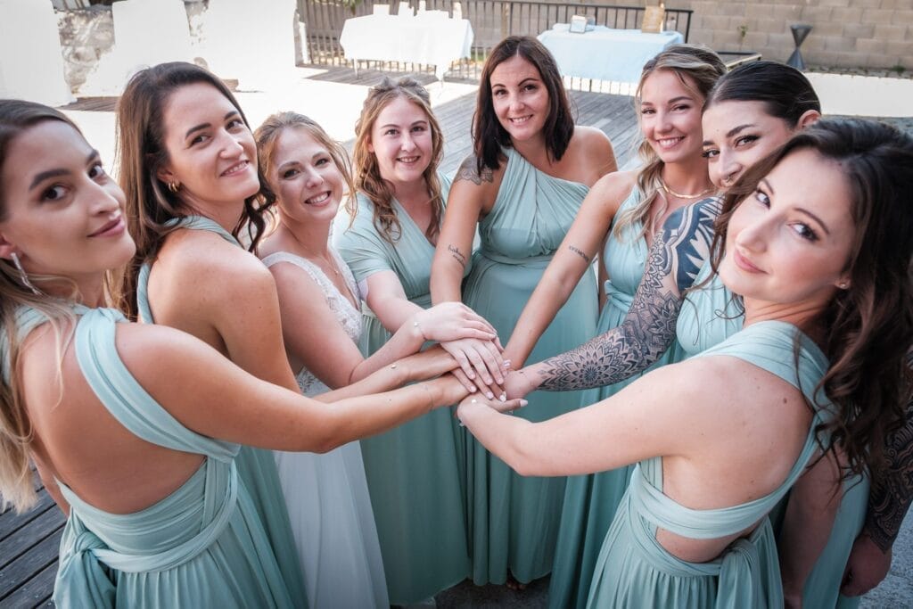 Hochzeitskrone Eine Gruppe von acht Frauen in passenden blaugrünen Kleidern steht lächelnd in einem Kreis, legt in der Mitte ihre Hände zusammen und fängt bezaubernde Momente ein, die natürliche Hochzeitsfotos wirklich ausmachen.