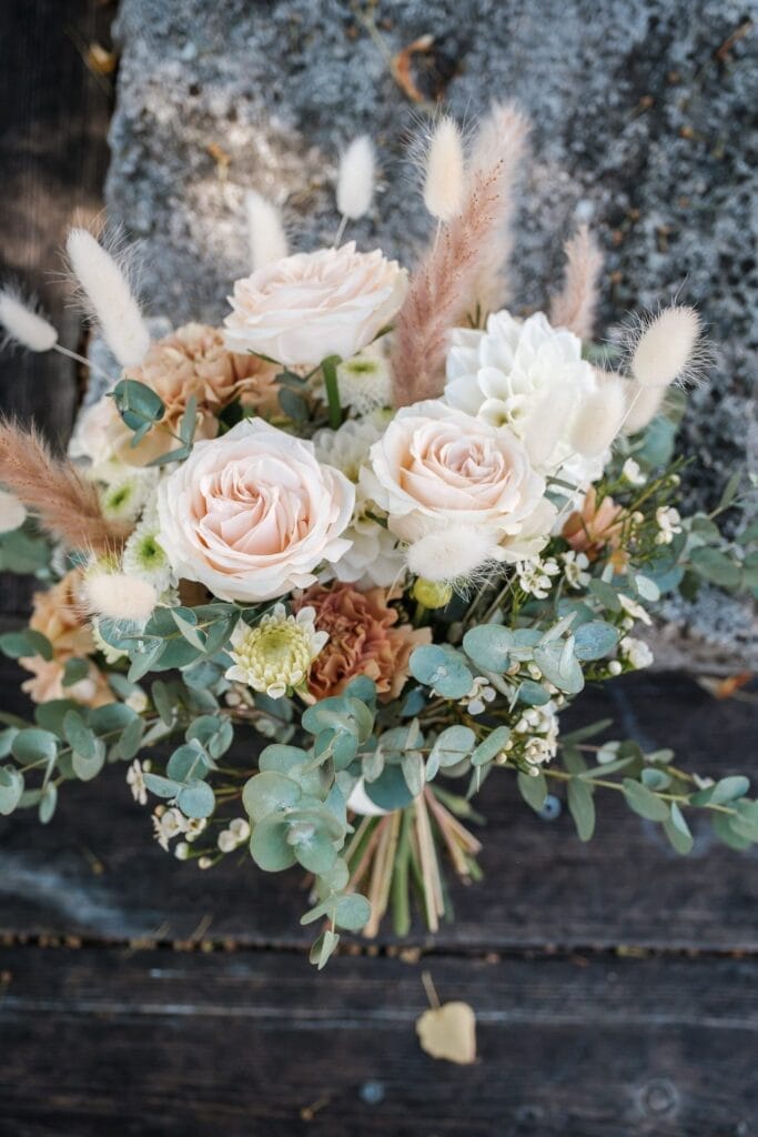 Hochzeitskrone Ein Strauß mit rosa Rosen, weißen Blumen, Eukalyptusblättern und weizenähnlichen Gräsern schmückt wunderschön einen strukturierten Stein- und Holzhintergrund und fängt bezaubernde Momente ein, perfekt für natürliche Hochzeitsfotos oder exquisite Hochzeitsfotografie in Niederösterreich.