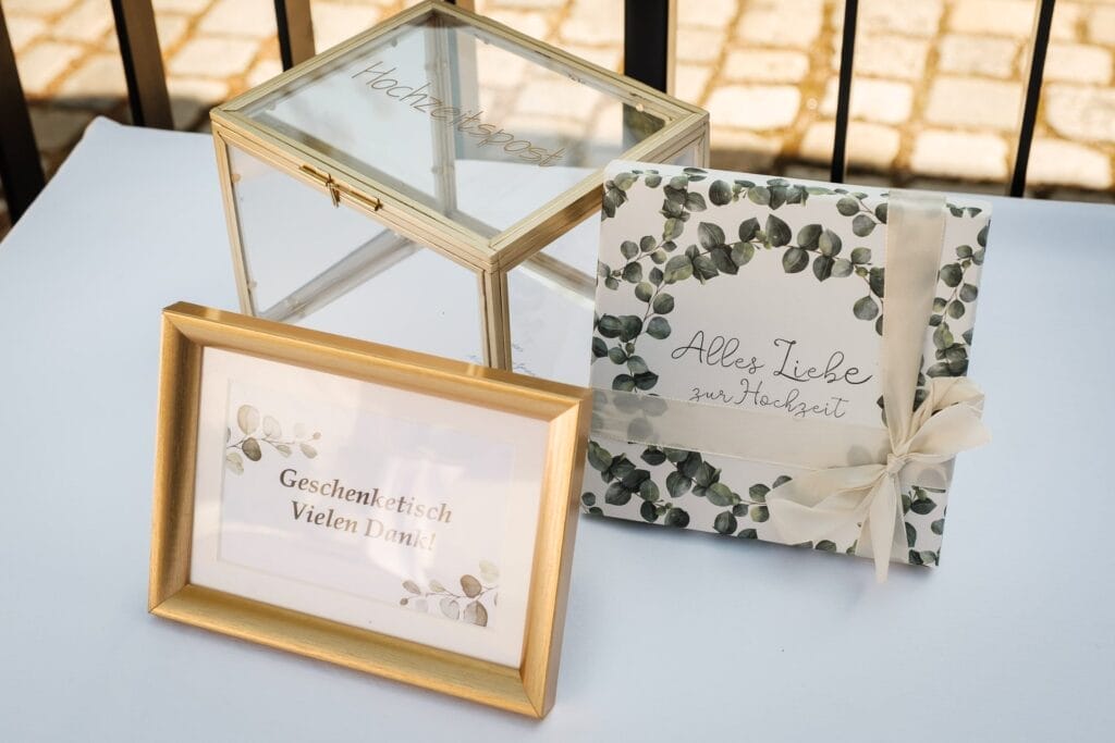 Hochzeitskrone Eine Glasbox mit der Aufschrift „Hochzeitspost“, ein gerahmtes Schild mit der Aufschrift „Geschenketisch Vielen Dank“ und ein verpacktes Geschenk mit der Aufschrift „Alles Liebe zur Hochzeit“ auf einem weißen Tisch fangen bezaubernde Momente ein, die die Schönheit der Hochzeitsfotografie in Niederösterreich hervorrufen.