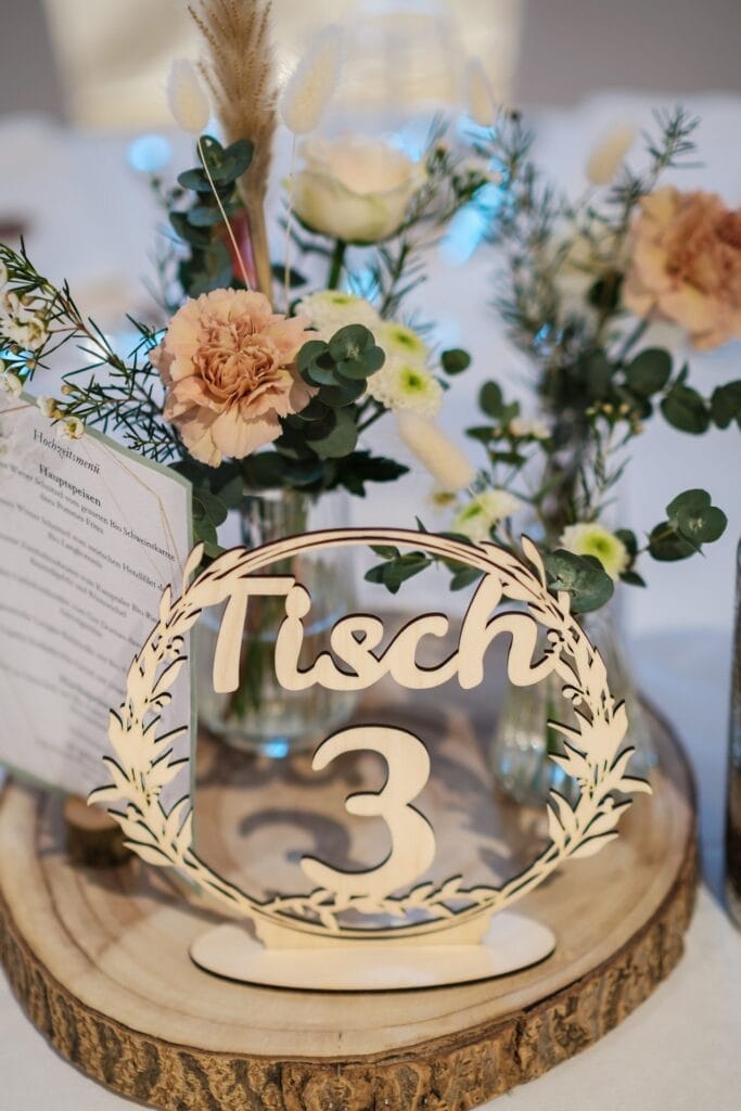 Hochzeitskrone Tischdekoration mit „Tisch 3“ in Holzbuchstaben auf einem geschnitzten Ständer, umgeben von Blumen und Grün auf einem Holzsockel, hält bezaubernde Momente fest. Eine Menükarte ziert den Hintergrund, perfekt für natürliche Hochzeitsfotos während Ihrer Liebesfeier.