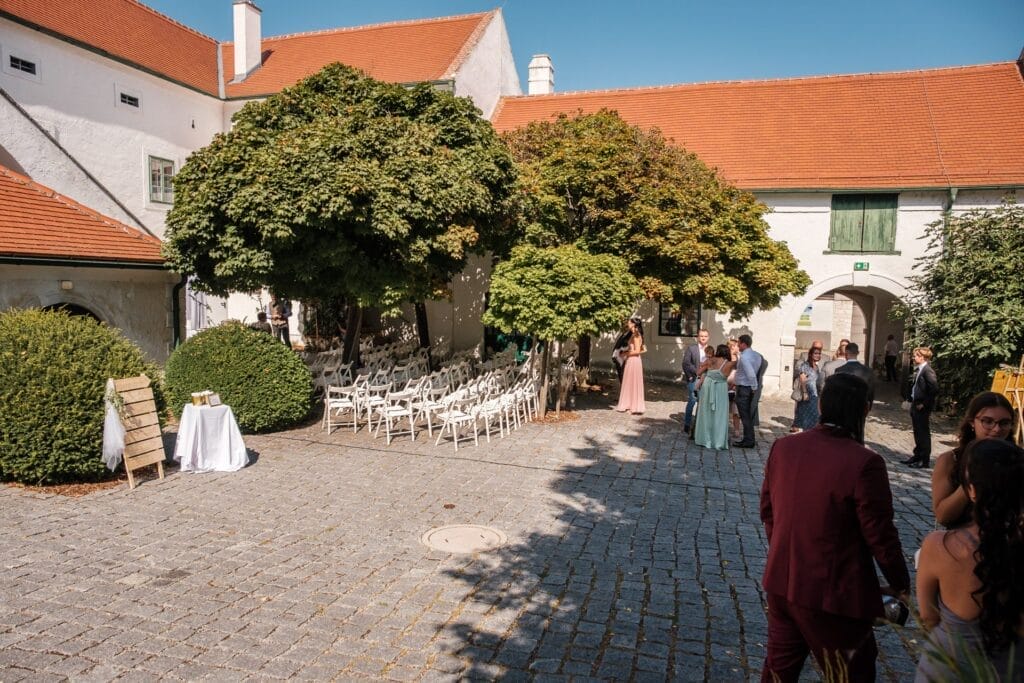 Hochzeitskrone In dieser malerischen Szene im Innenhof mischen sich die Menschen unter den üppigen Bäumen, während Reihen weißer Stühle auf die Gäste warten. Die charmanten Gebäude mit den roten Dächern, die den Bereich umgeben, bilden eine perfekte Kulisse für bezaubernde Momente und fangen natürliche Hochzeitsfotos in Niederösterreich unter einem klaren Himmel ein.