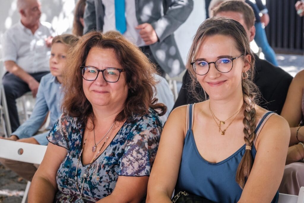 Hochzeitskrone Zwei Frauen mit Brillen lächeln und fangen bezaubernde Momente ein. Hinter ihnen sorgen ein kleiner Junge und andere in formeller Kleidung für den Charme – ein exquisiter Hauch von Hochzeitsfotografie in Niederösterreich.