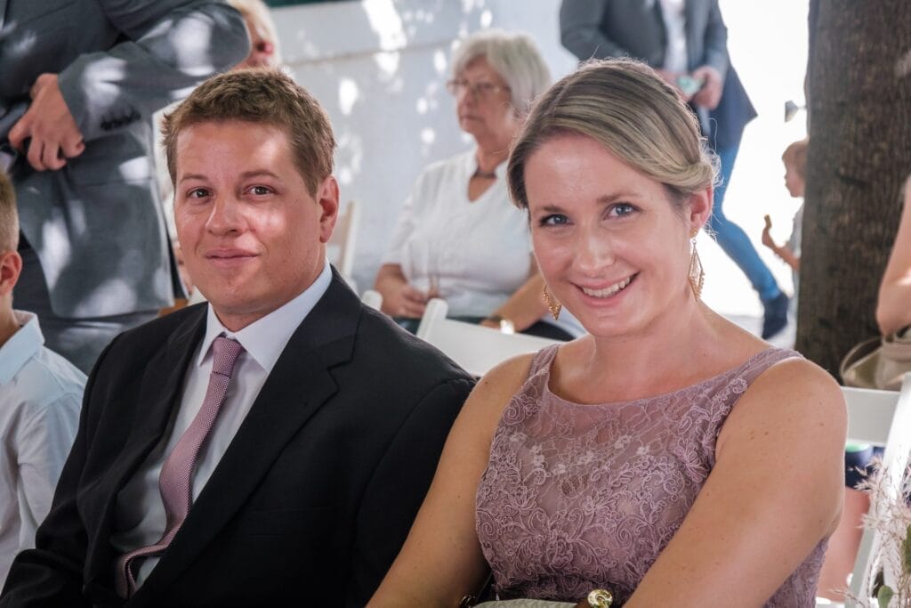 Hochzeitskrone Ein Mann im Anzug und eine Frau im Spitzenkleid sitzen lächelnd bei einer formellen Veranstaltung und fangen bezaubernde Momente ein, die an Hochzeitsfotografie in Niederösterreich erinnern, während sich im Hintergrund die anderen Gäste unter die Leute mischen.