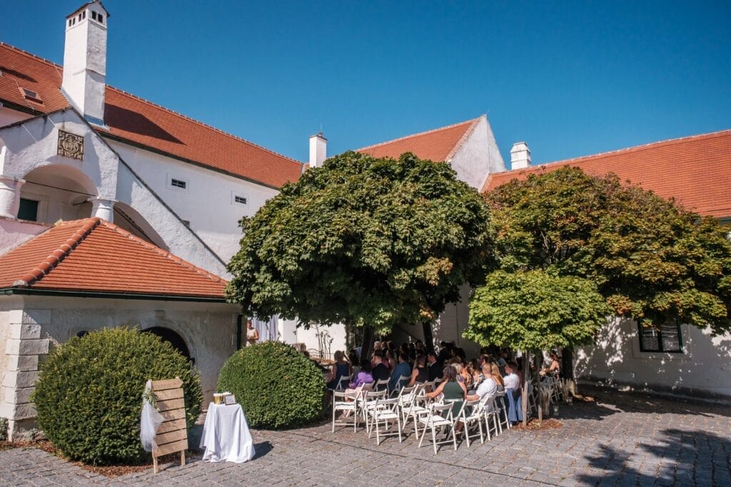 Hochzeitskrone Ein bezauberndes Treffen im Freien mit sitzenden Teilnehmern findet unter großen Bäumen neben einem weißen Gebäude mit roten Dächern statt und fängt bezaubernde Momente an einem sonnigen Tag ein. Perfekt für diejenigen, die natürliche Hochzeitsfotos suchen oder Hochzeitsfotografie in Niederösterreich erkunden möchten.