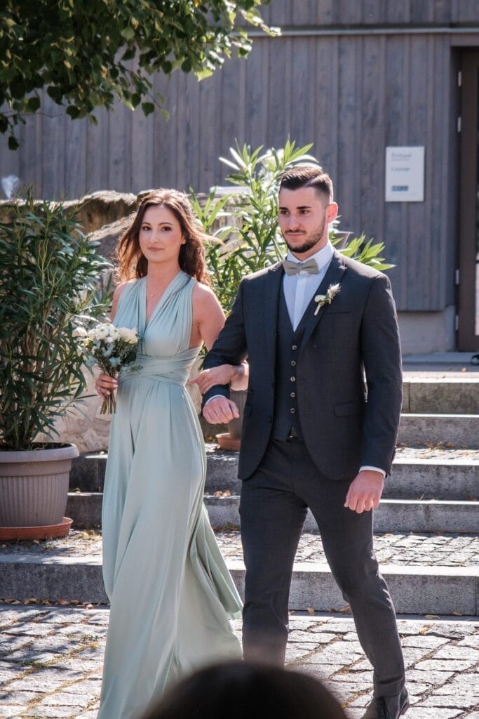 Hochzeitskrone Eine Frau in einem hellgrünen Kleid hält Blumen in der Hand und geht Arm in Arm mit einem Mann im dunklen Anzug und fängt bezaubernde Momente im Freien an einem sonnigen Tag ein.